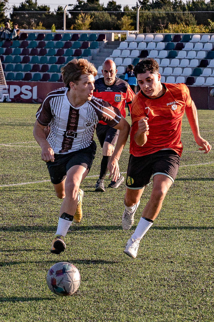 Dos jugadores corriendo hacia la pelota en el césped, con tribuna detrás
