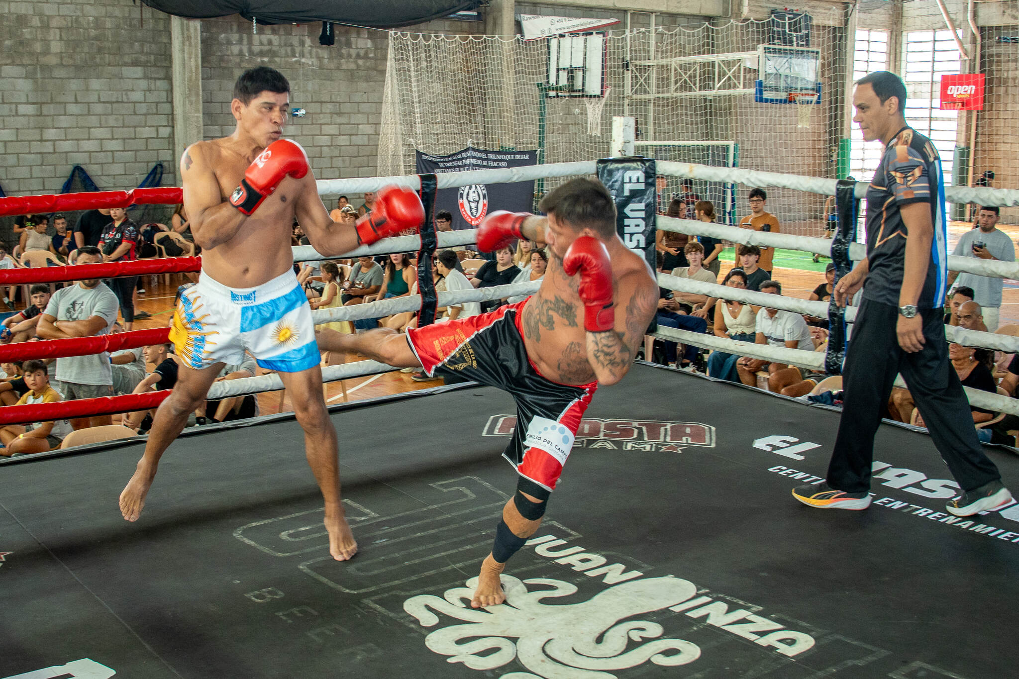 Patada en pleno combate sobre el ring, con el árbitro al costado y el público alrededor